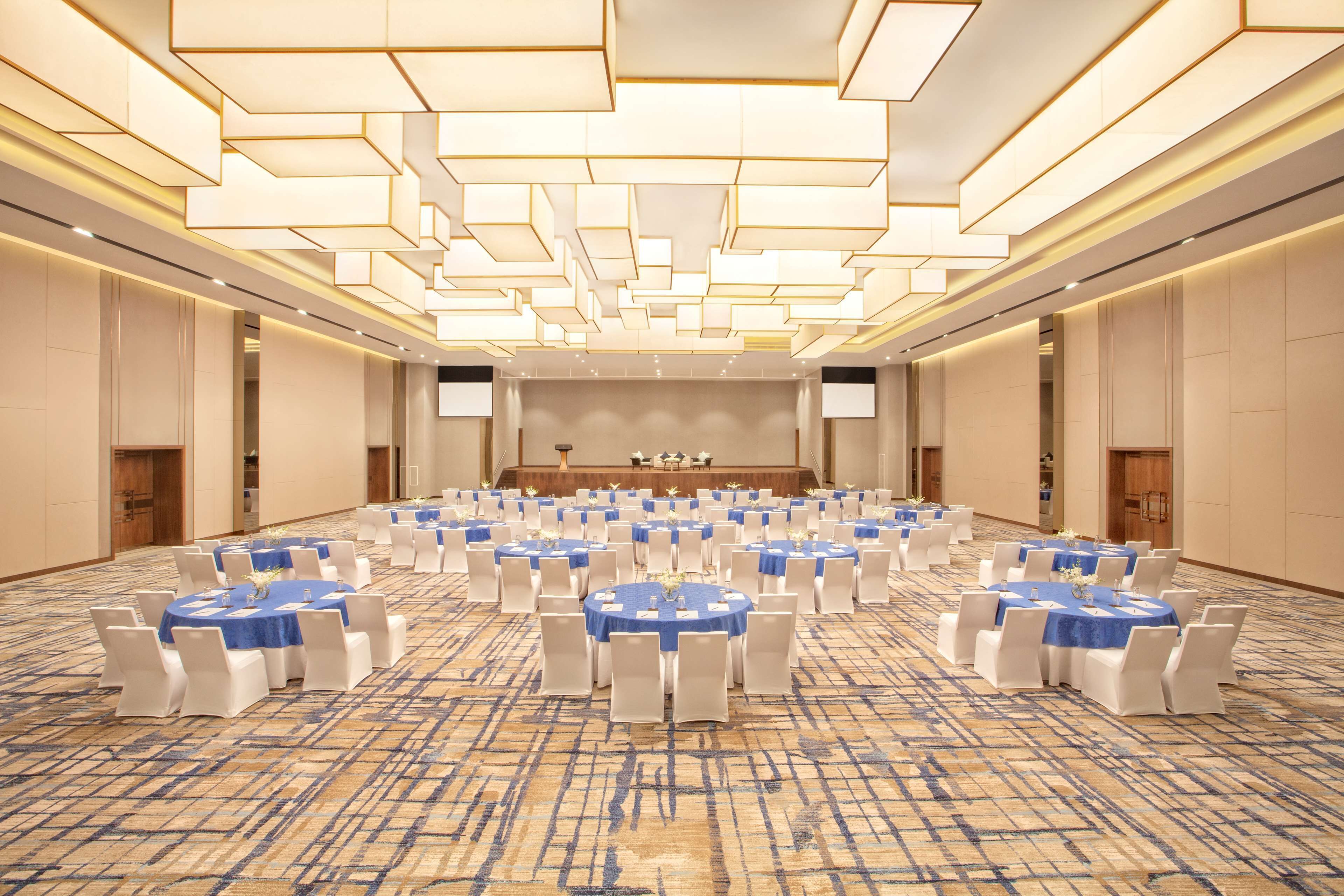 A large conference room with multiple round tables, each with white chairs and blue tablecloths. The room features a patterned carpet and a modern ceiling design with recessed lighting.