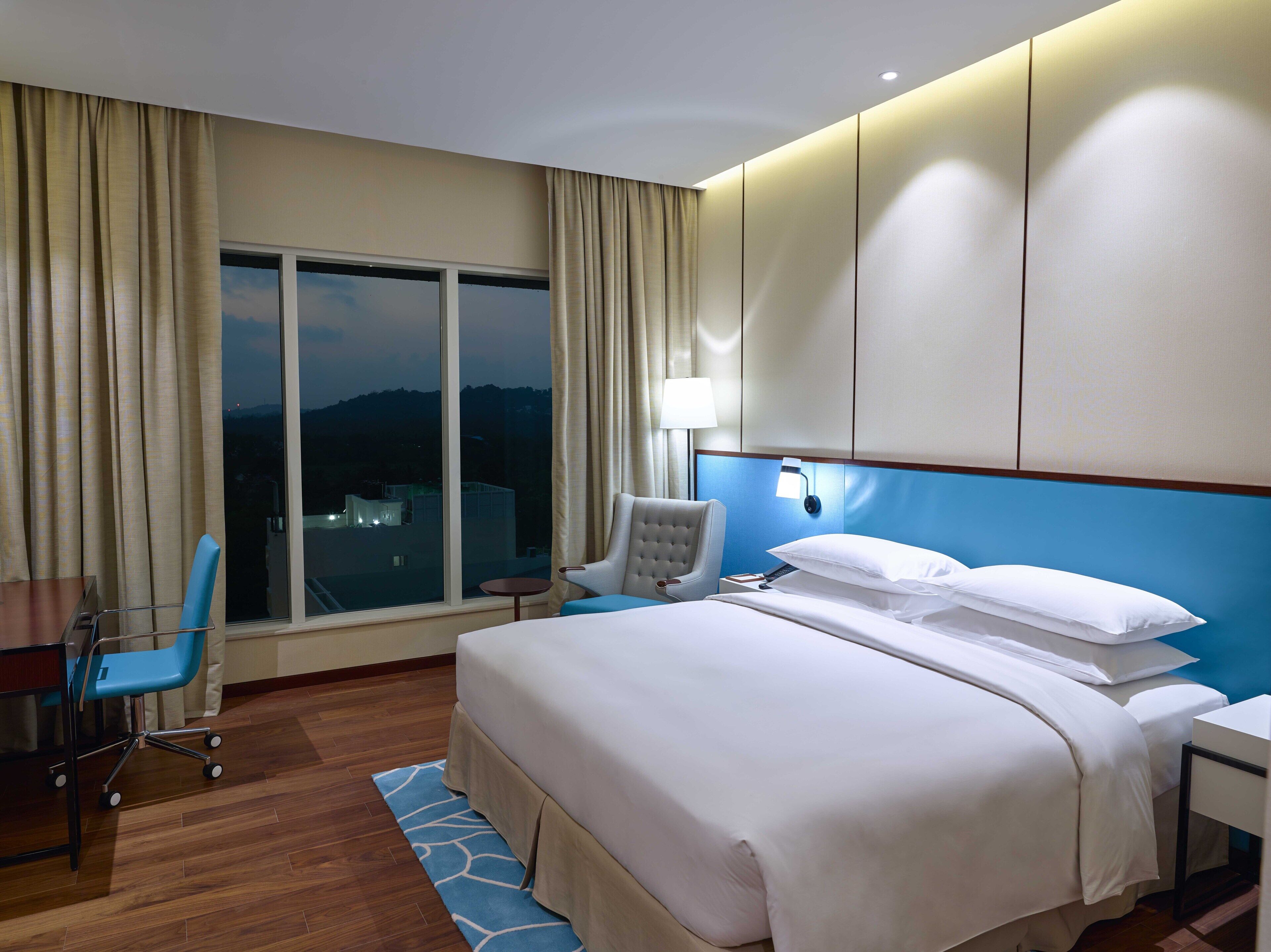 A hotel room with a large bed, a desk with a chair, a window with curtains, and a view of the city.