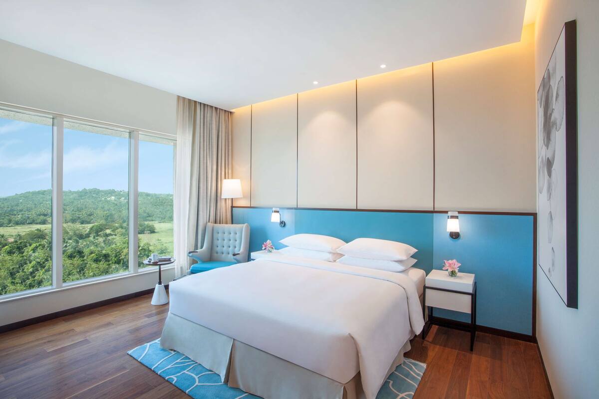 Una habitación de hotel moderna con una cama grande, una mesita de noche, una lámpara y vistas al exterior.