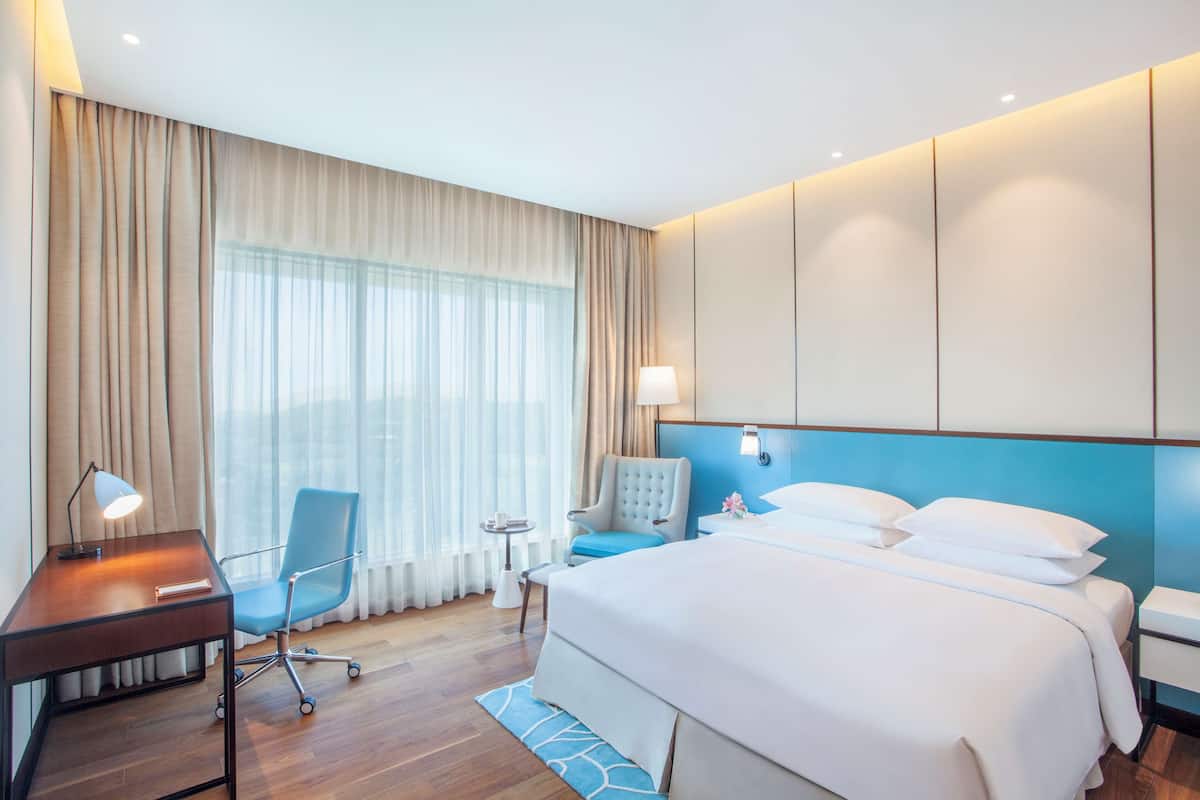 Habitación de hotel con una cama grande, un escritorio con lámpara, una silla, una mesita y una ventana con cortinas.