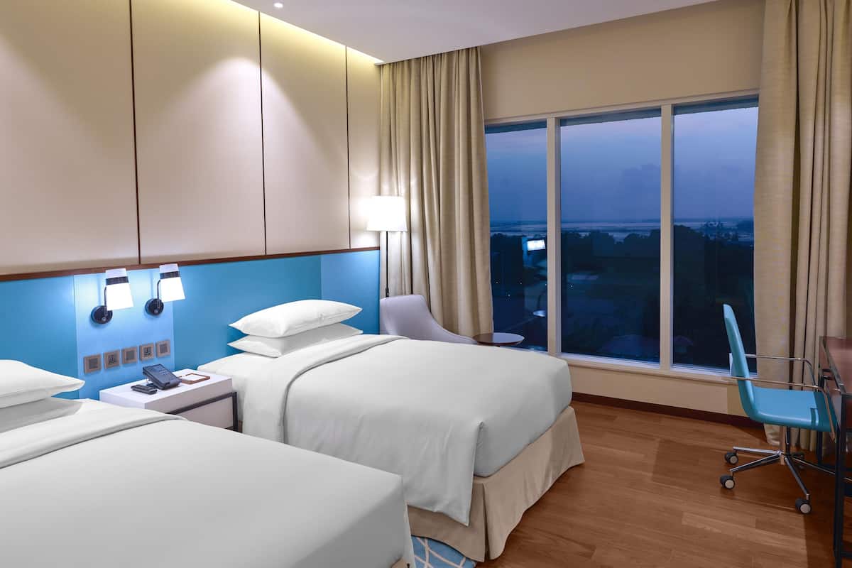 Habitación de hotel con dos camas, un escritorio, una silla, una lámpara y un amplio ventanal con vistas.