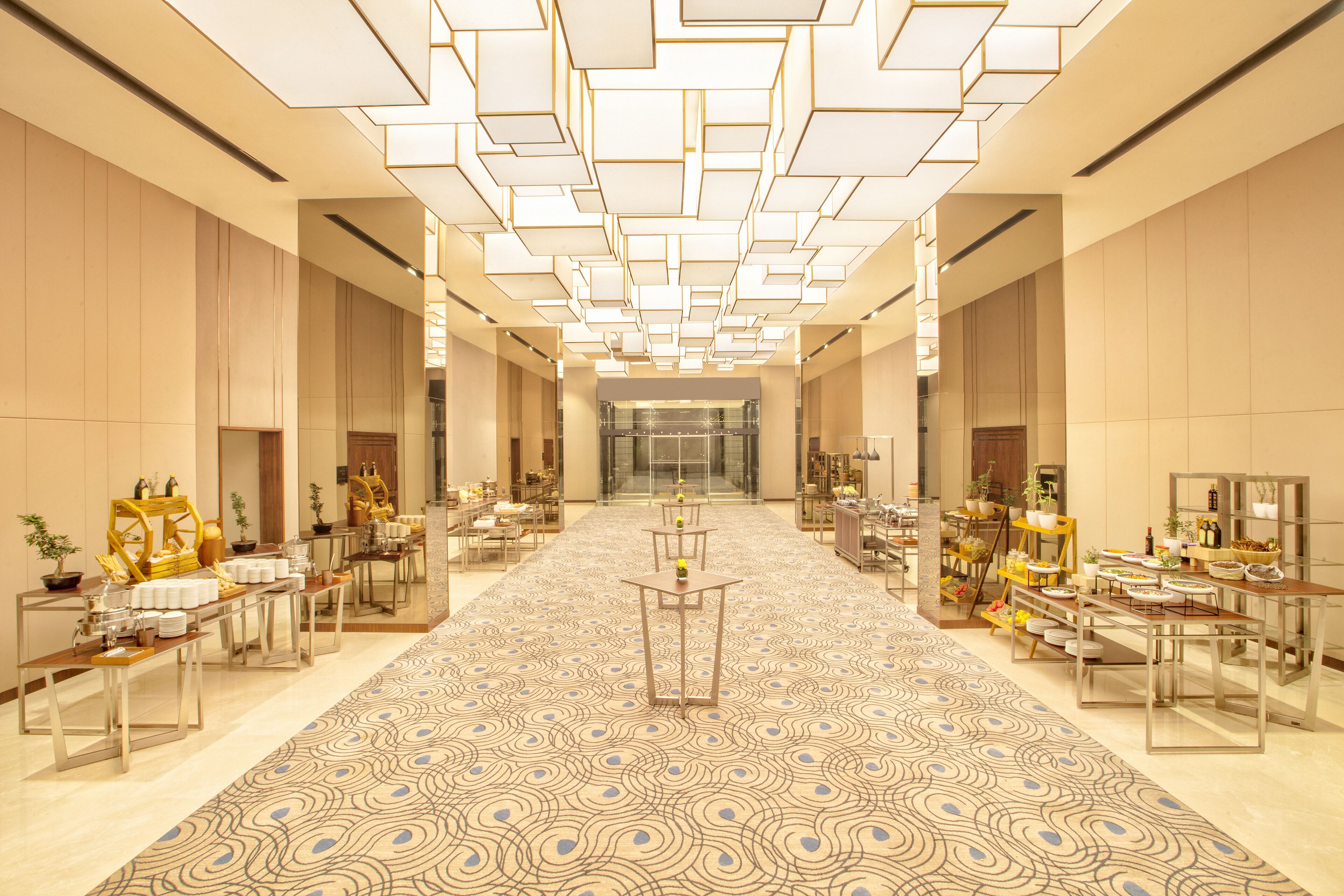 A spacious hotel lobby with a patterned carpet, multiple tables with amenities, and a modern ceiling design.