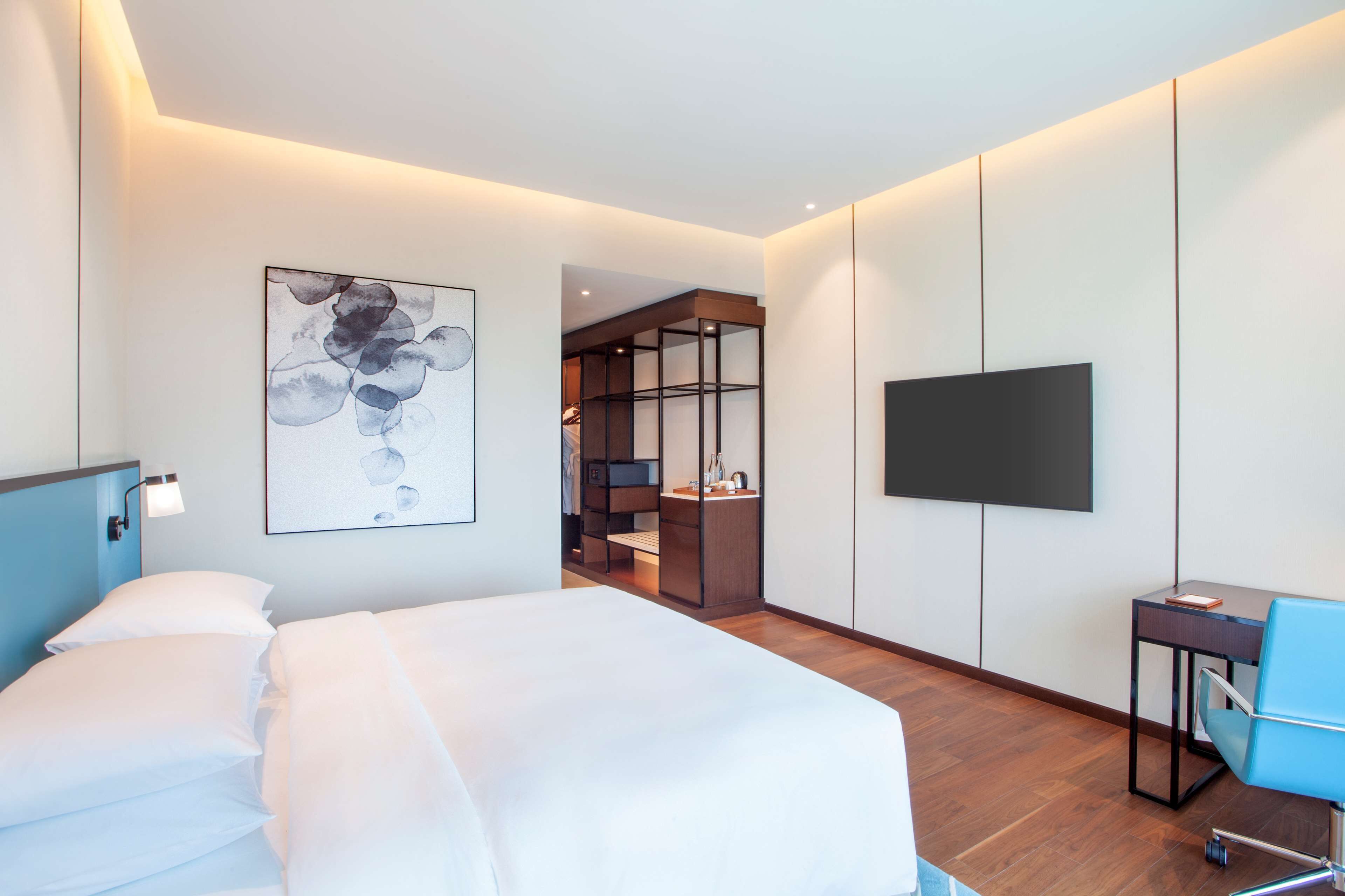 A modern hotel room with a large bed, a desk with a chair, a flat-screen TV, and a bookshelf.