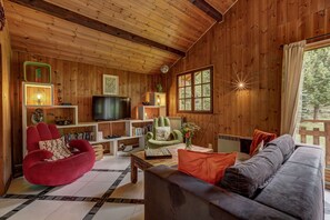 A cozy living room with wooden walls, a red armchair, a green armchair, a grey sofa, and a television on a wooden shelf.