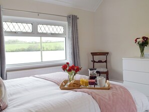 Una camera da letto con un letto, una finestra vista su un campo verde, una sedia, un comò e un vassoio con cose per la colazione.