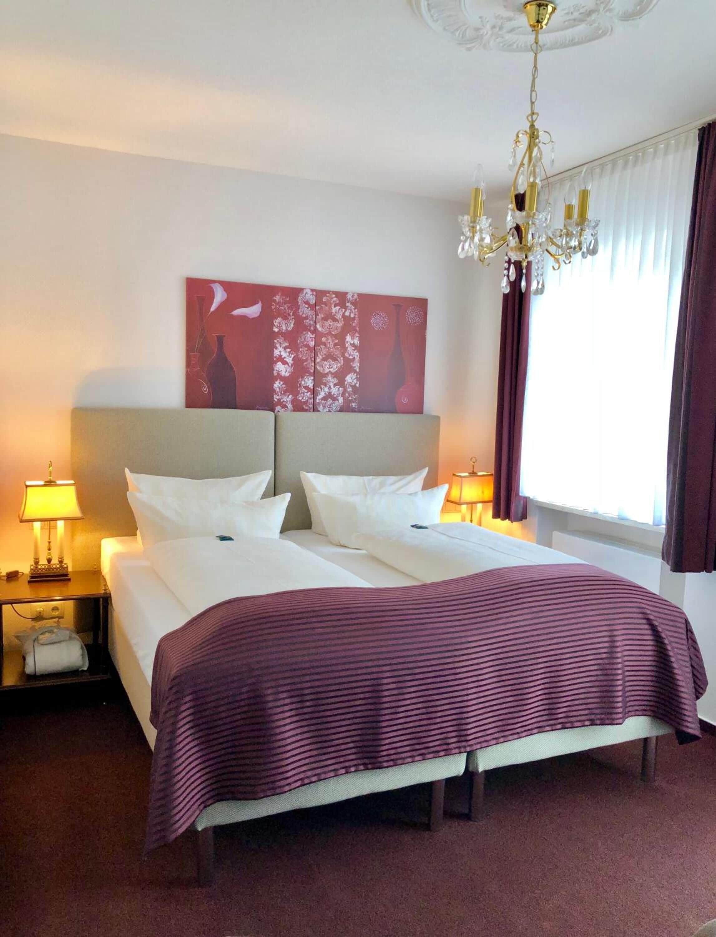 A hotel room with a large bed, two bedside lamps, a chandelier, a painting on the wall, and a window with curtains.