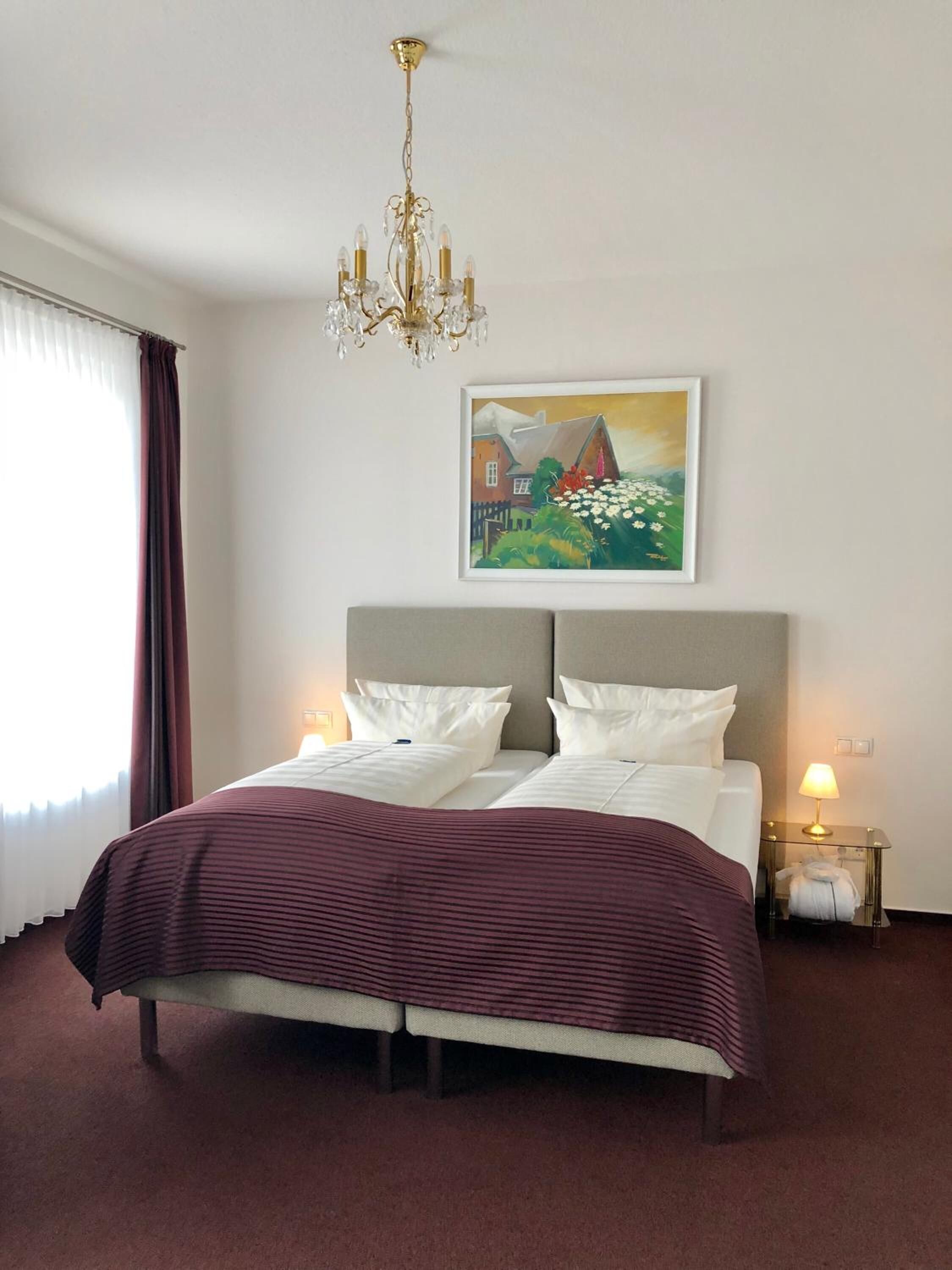 A hotel room with a large bed, a chandelier, a painting on the wall, and a window with curtains.