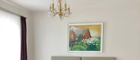A hotel room with a large bed, a chandelier, a painting on the wall, and a window with curtains.