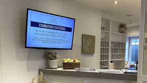 A hotel reception area with a television displaying a message in German, a potted plant, a basket of fruits, and a decorative wall piece. - Hotel Sylter Blaumuschel (Sylt)