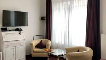 A modern hotel room with a flat-screen TV, a minibar, a sofa, a chair, and a glass coffee table.