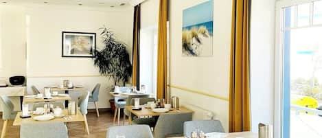 Uma sala de jantar de hotel com várias mesas e cadeiras, uma grande janela com cortinas e obras de arte nas paredes.