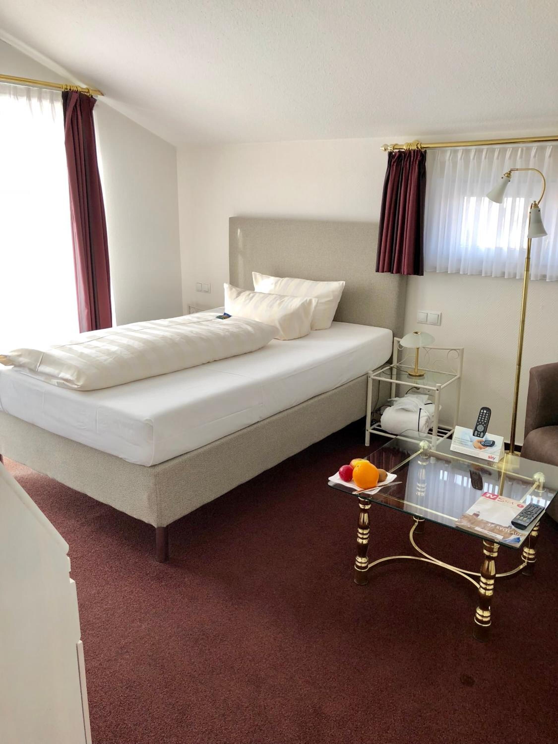 Une chambre d’hôtel avec un lit, une table basse en verre avec une télécommande, une lampe et une petite table sur laquelle sont disposés des fruits.