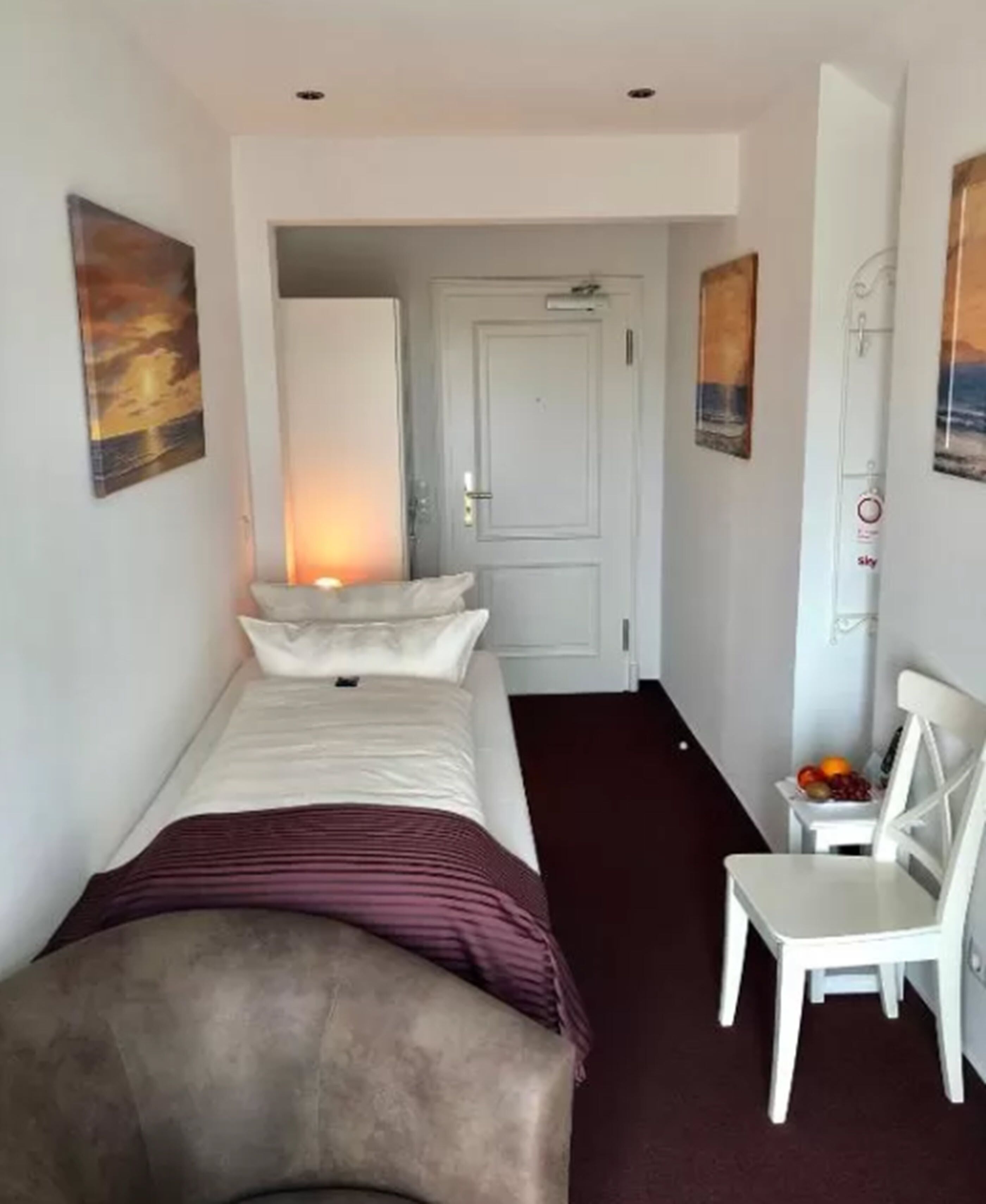 Ein schmales Hotelzimmer mit einem Einzelbett, einem Sessel und einem kleinen Tisch mit Obst.
