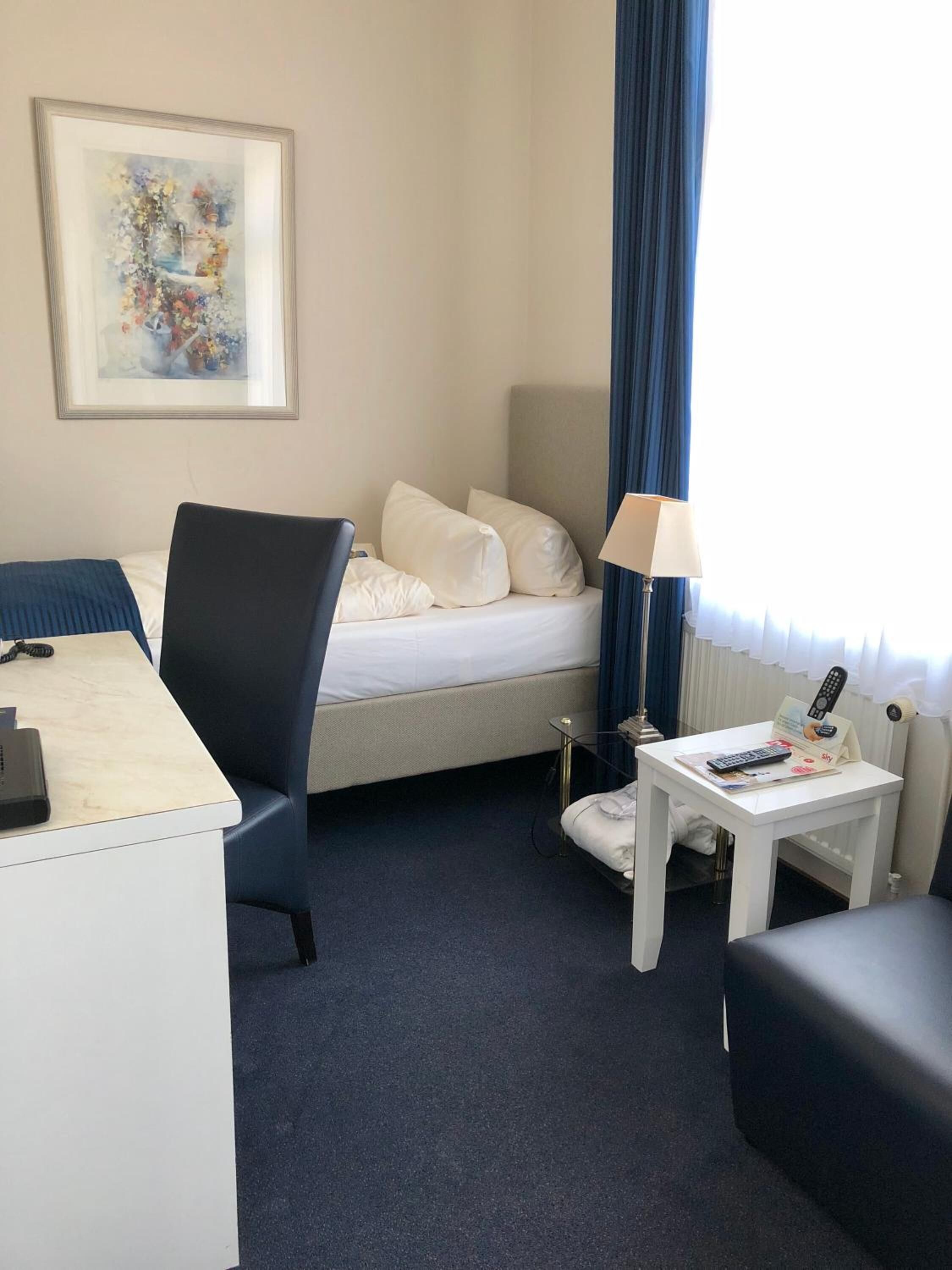 Ein Hotelzimmer mit Bett, Schreibtisch, Stuhl, kleinem Tisch mit Lampe und einem Bild an der Wand.