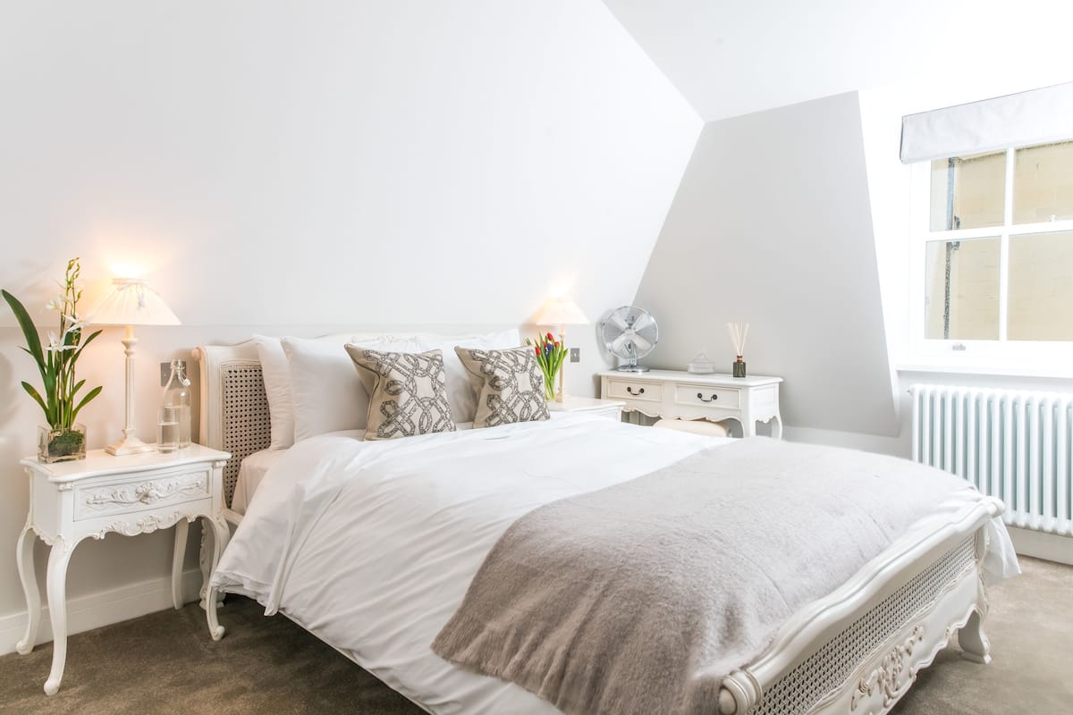 A bedroom with a large bed, white nightstands, a dresser, a window with blinds, and a radiator.