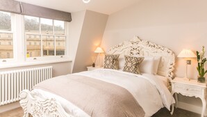 Un dormitorio con una cama grande, cabecera blanca ornamentada, dos mesitas de noche con lámparas, una ventana con vistas a edificios y un radiador.