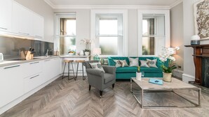 Una sala de estar moderna con un sofá azul verdoso, un sillón gris y una mesa de centro. Hay una zona de cocina con armarios blancos y un suelo con patrón de espiga.