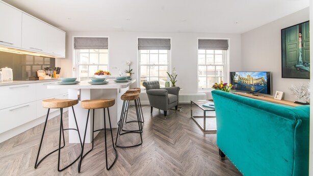 Una cocina moderna con armarios blancos, una zona de comedor con un sofá color turquesa y un televisor sobre una mesa de madera.