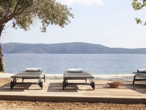 A wooden deck with three sun loungers facing a body of water, a tree to the left, and a distant mountain range. - Ilya Suites in Alonnisos, Sporades Islands, Greek Islands, (Steni Vala)
