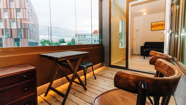 A balcony with wooden furniture, a view of a modern building, and a sliding glass door leading to an interior room. - Tower Apartments (Reykjavik)