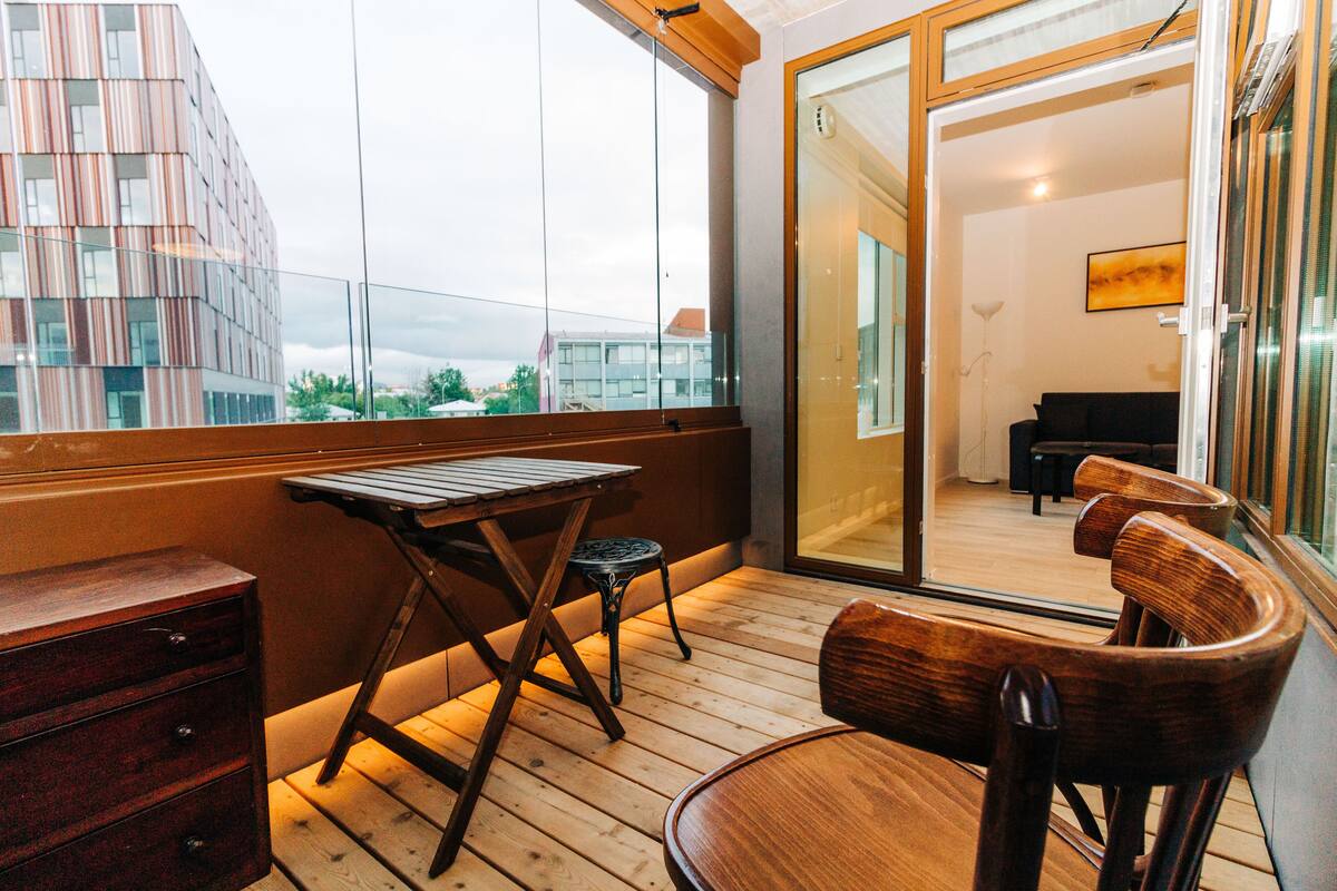 A balcony with wooden furniture, a view of a modern building, and a sliding glass door leading to an interior room.