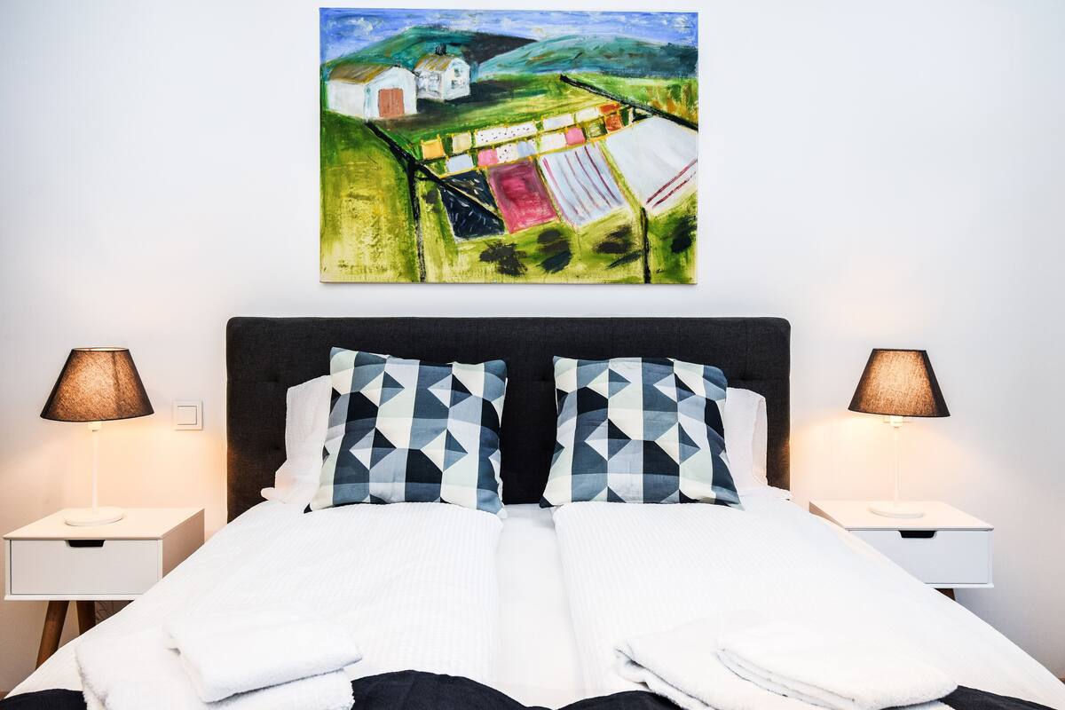 A neatly made bed with a geometric patterned pillow, two bedside lamps, and a painting of a rural landscape above the bed.