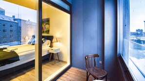 A modern hotel room with a large window, a bed, a bedside table, and a chair. - Tower Apartments (Reykjavik)