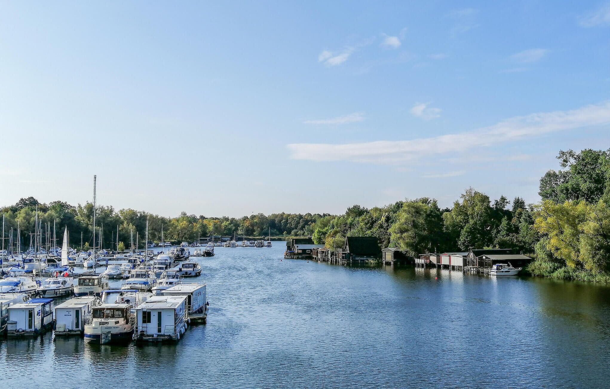 Un port de plaisance où sont amarrés de nombreux bateaux, ainsi qu’une rivière bordée de bâtiments et de verdure.