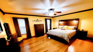 Une chambre avec un plancher en bois, un lit avec des oreillers, une commode, un téléviseur et un ventilateur de plafond.
