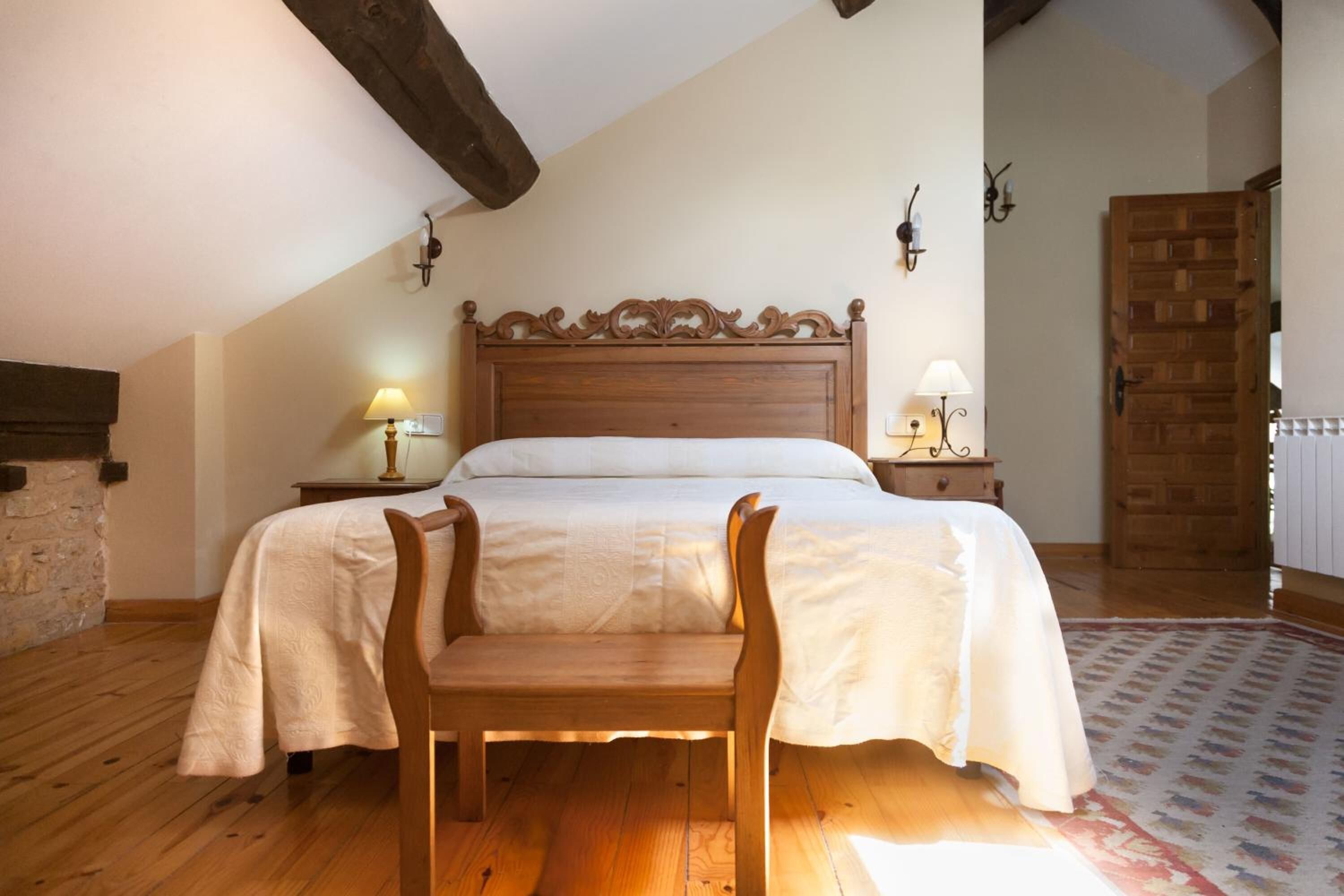 A bedroom with a wooden bed, two bedside tables with lamps, a stone wall, and a wooden door.