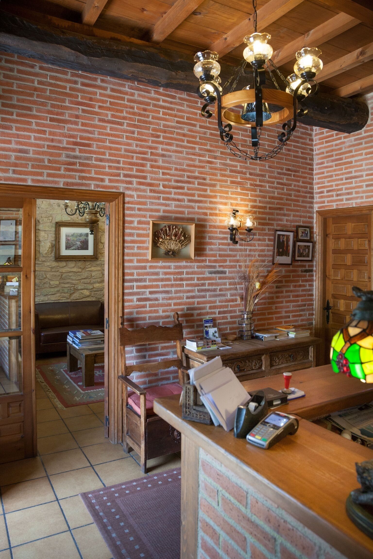 Un interior rústico con paredes de ladrillo visto, techo de madera, una lámpara de araña, mostrador de recepción y una zona de estar con sofá y mesa.
