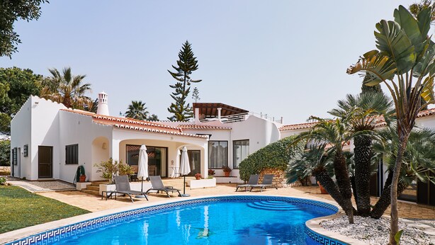 Un espace aménagé au bord de la piscine, avec un bâtiment blanc, un espace salon couvert et un palmier.