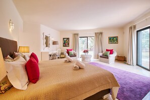 A spacious bedroom with a large bed, two chairs, a small table, and a view of the outdoors through large windows. - Villa Shambala (Carvoeiro)