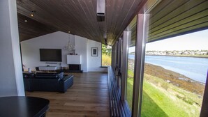 Una sala de estar moderna con un televisor grande, chimenea y sofá. Cuenta con piso de madera y ventanas amplias con vistas a un cuerpo de agua.