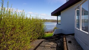Un edificio moderno con vistas a un lago, un sendero de grava que lleva a la entrada y un área ajardinada con arbustos.