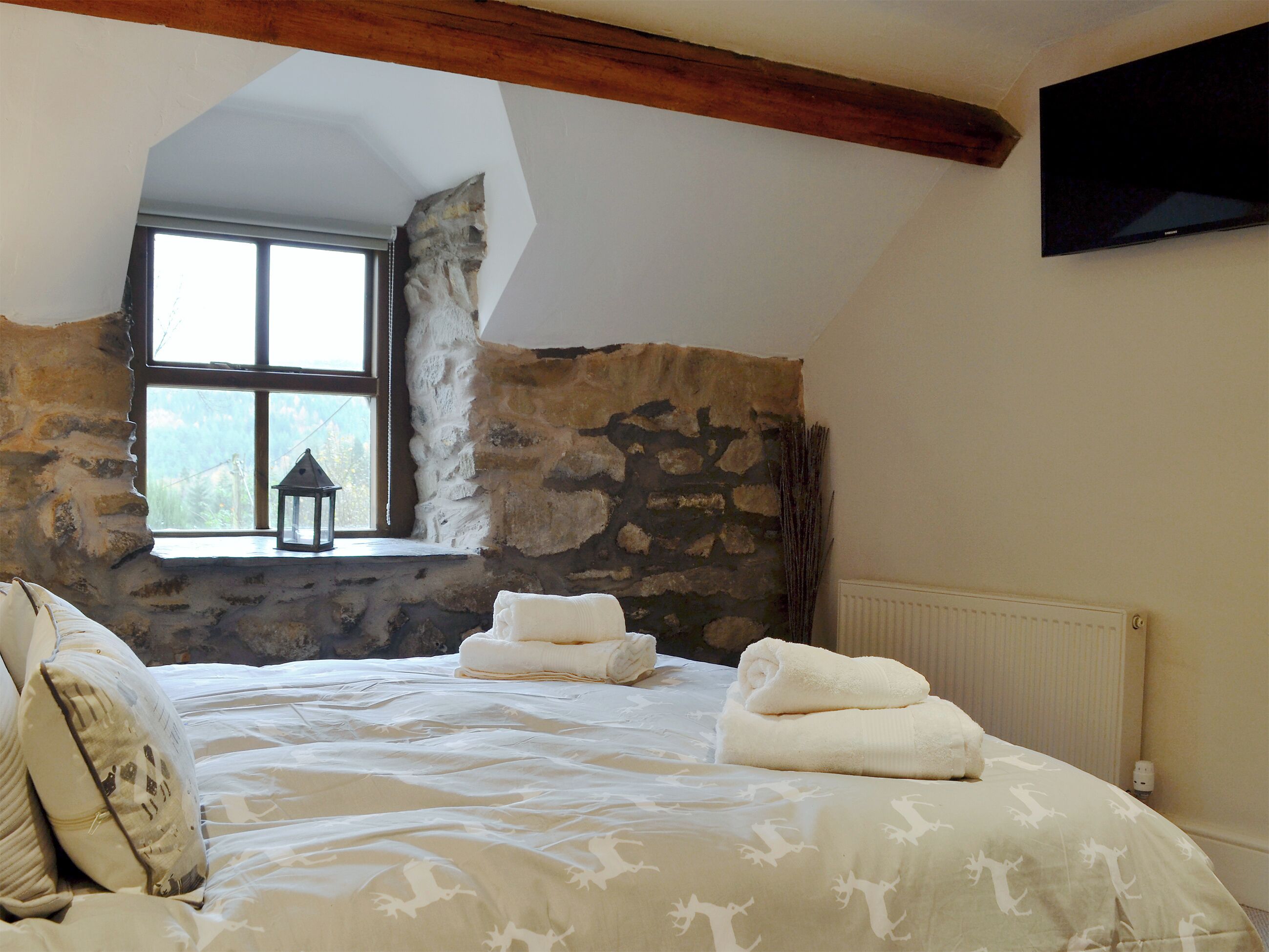 A bedroom with a stone wall, a window with a lantern, a bed with white bedding, and a flat-screen TV mounted on the wall.