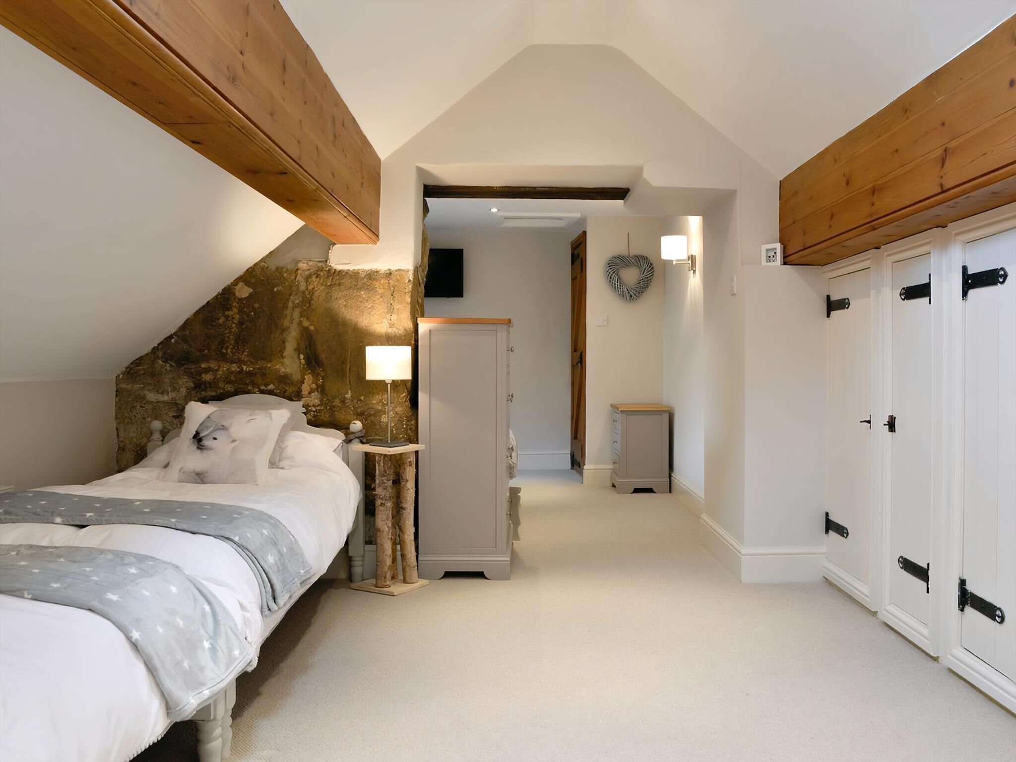 A bedroom with a sloped ceiling, a bed with a star-patterned blanket, a bedside table with a lamp, a wardrobe with black handles, and a hallway leading to another room.