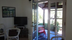 Un salon équipé d’un téléviseur, de fauteuils en osier, d’une table rouge et d’une porte vitrée donnant sur une terrasse extérieure avec un fauteuil rouge et de la verdure.