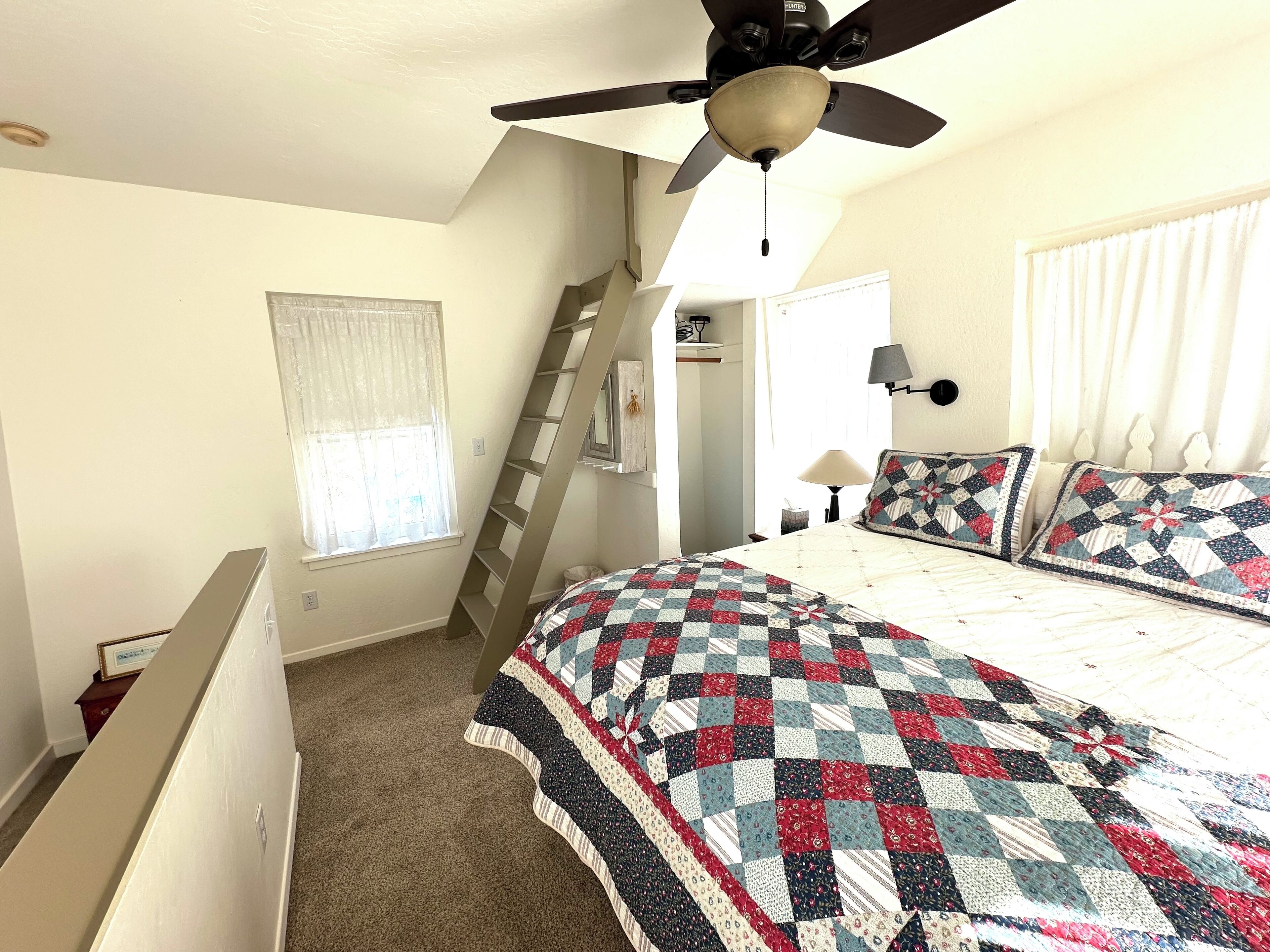 Une chambre à coucher avec un lit, un ventilateur de plafond, une fenêtre avec des rideaux, une échelle et une lampe de chevet.