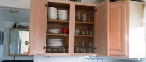 A kitchen with open cabinets containing dishes, glasses, and a sink with a mirror above it.