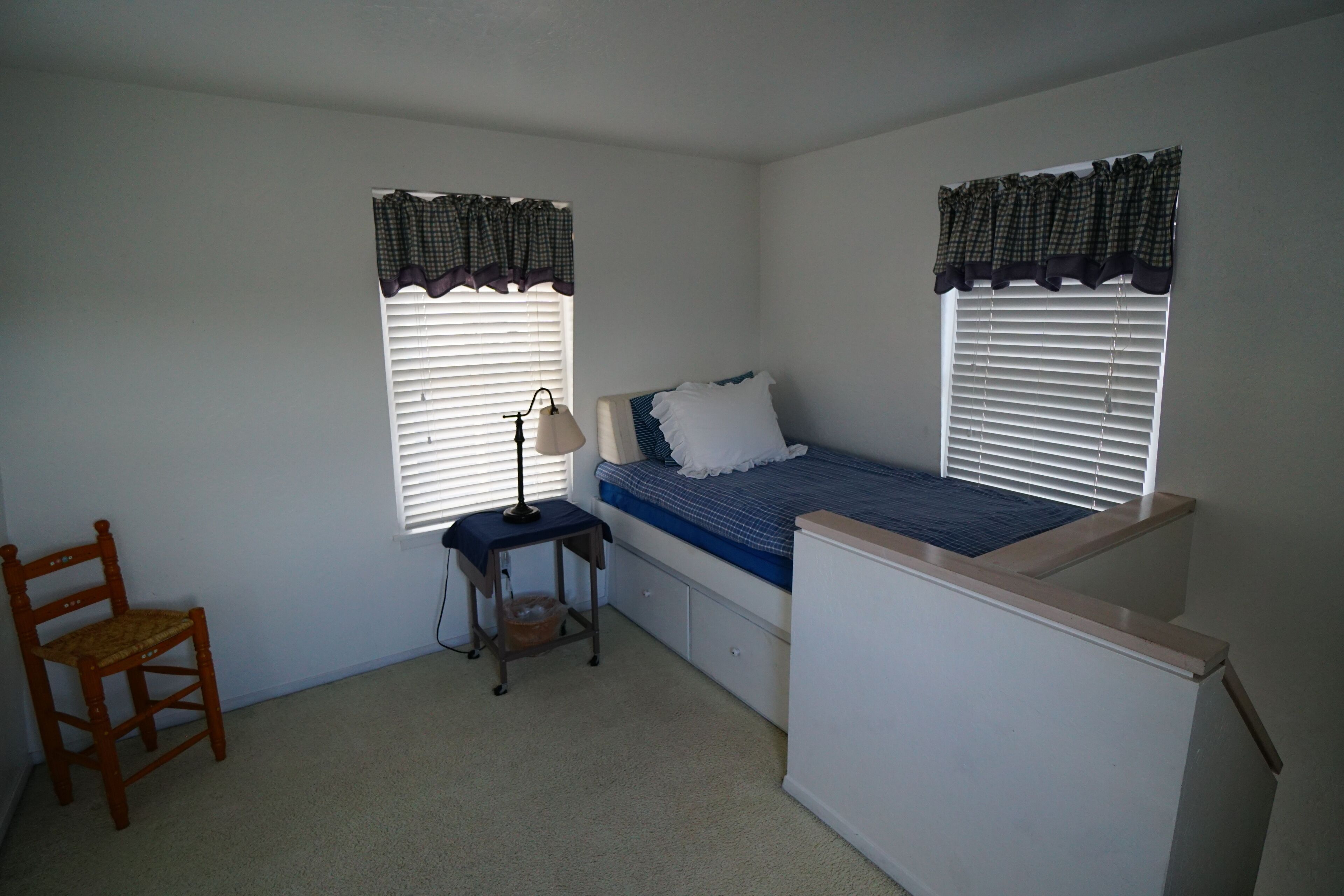 A bedroom with a single bed, two windows with curtains, a chair, a bedside table with a lamp, and a carpeted floor.