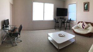 Una sala de estar con un sofá blanco, una mesa de centro blanca, un televisor y un escritorio con una computadora.