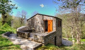 Una cabaña de piedra con puerta de madera, un pequeño porche y un camino de piedra que la conduce.