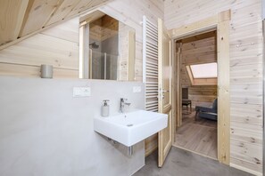 Un bagno moderno con lavandino, specchio e doccia bianchi. Sono visibili pareti in legno e un soffitto spiovente con finestra a lucernario.