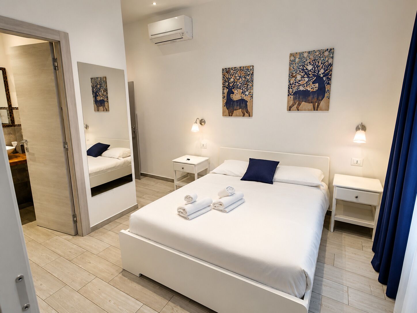 Moderni hotellihuone, jossa on suuri sänky, kaksi yöpöytää, peili ja taideteoksia seinillä.