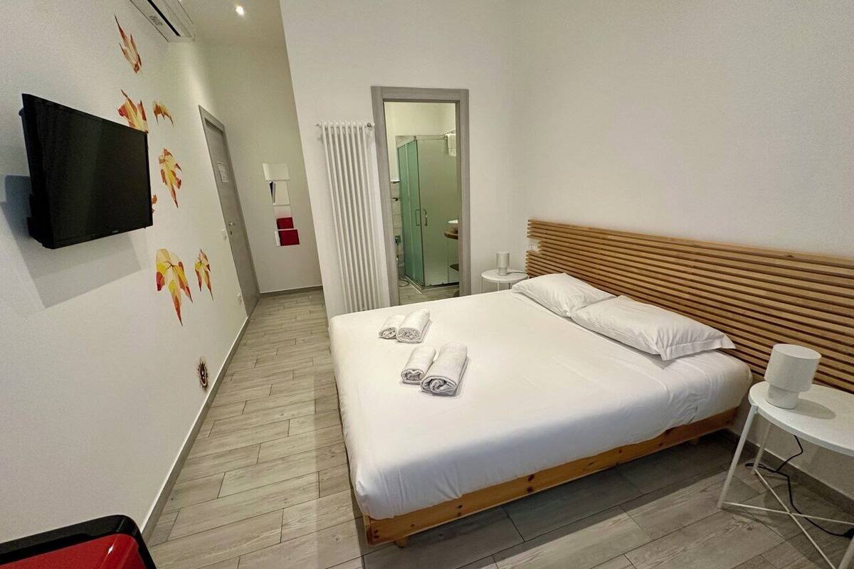 Moderni hotellihuone, jossa on puinen sänky, valkoiset vuodevaatteet, litteänäyttöinen televisio ja pieni valkoinen pöytä.