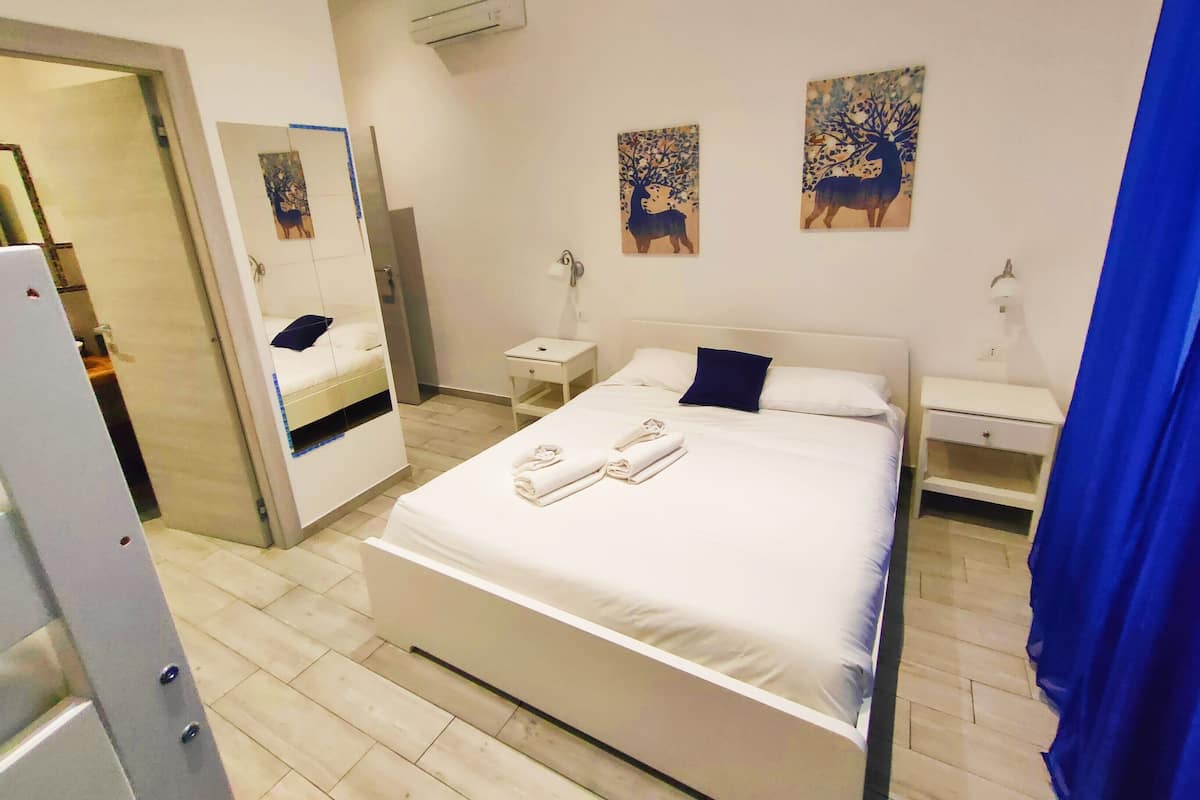 Hotellihuone, jossa on sänky, yöpöytä, peili ja kaksi taulua seinällä.