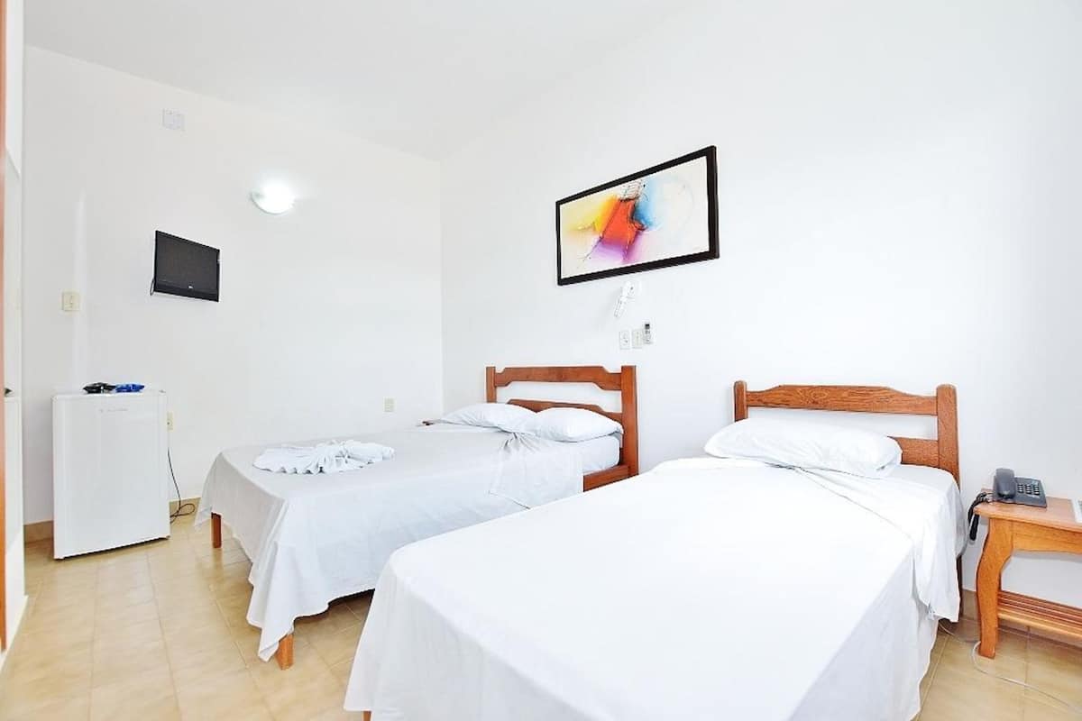 Quarto de hotel com duas camas de solteiro, uma secretária pequena com telefone, televisão fixada na parede e um frigorífico.