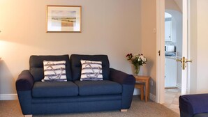 A living room with a dark blue sofa, two pillows with anchor designs, a framed landscape painting, a small table with a vase of flowers, and a doorway leading to a kitchen. - 3 bedroom accommodation in Brancaster Staithe, near Wells-next-the-Sea (Brancaster Staithe, near Wells-next-the-Sea)