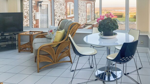 A conservatory with a TV, wicker furniture, a round table with a potted plant, and a view of a brick building and landscape. - 3 bedroom accommodation in Brancaster Staithe, near Wells-next-the-Sea (Brancaster Staithe, near Wells-next-the-Sea)